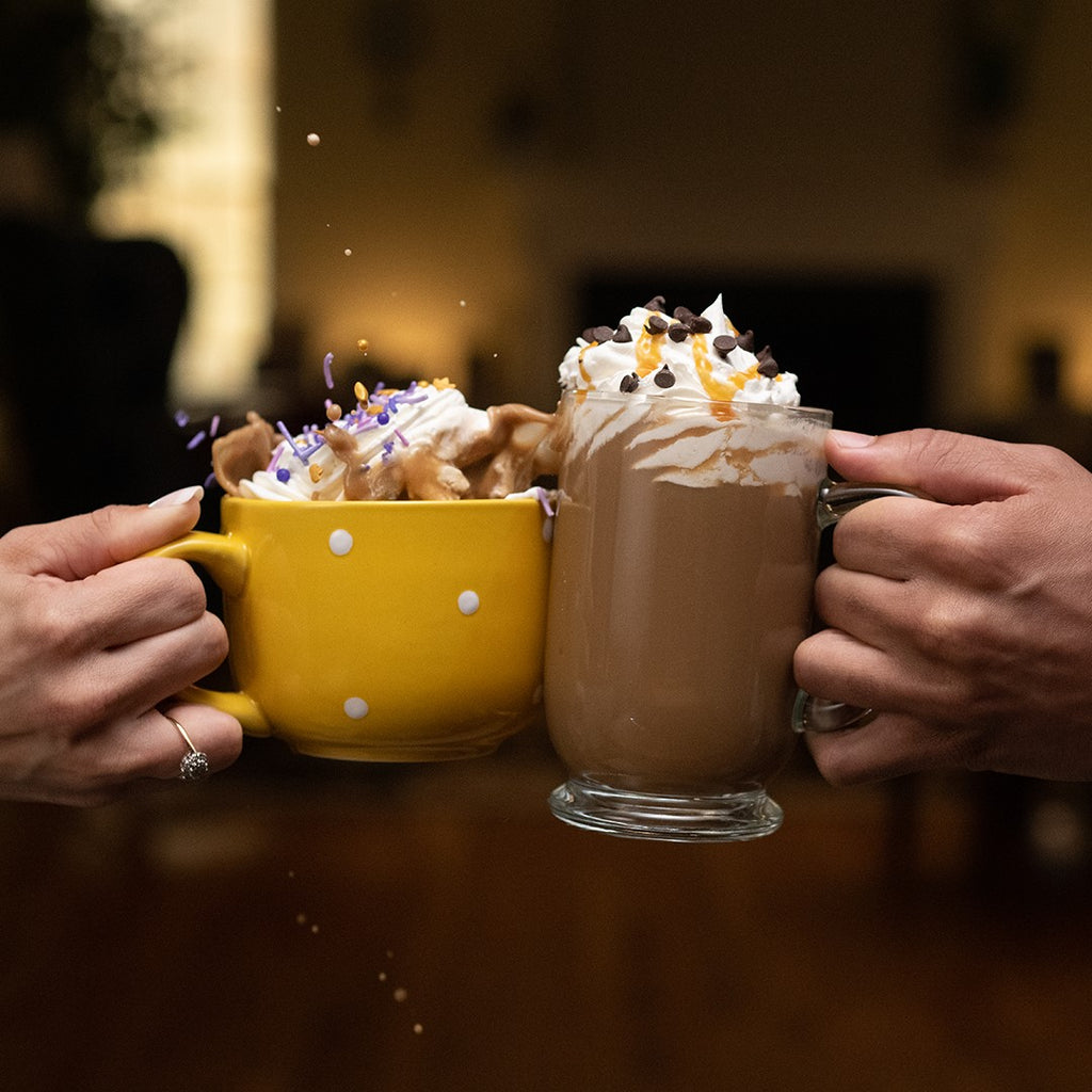 Two people holding a yellow mug of Hills Bros. Cappuccino Instant Cappuccino Mix - Sugar Cookie and a glass of hot chocolate topped with whipped cream and sprinkles, in a cozy, dimly lit setting.