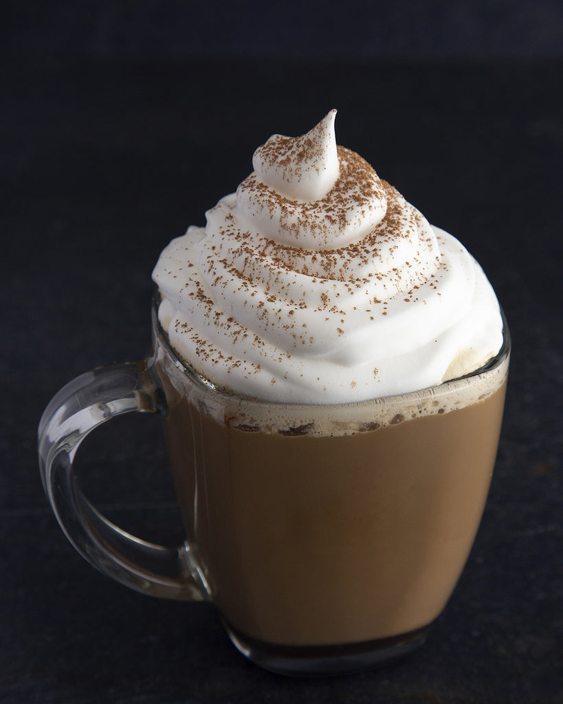 A clear glass mug containing a hot, low-calorie Hills Bros. Cappuccino topped with whipped cream and a dusting of spice, on a dark surface.