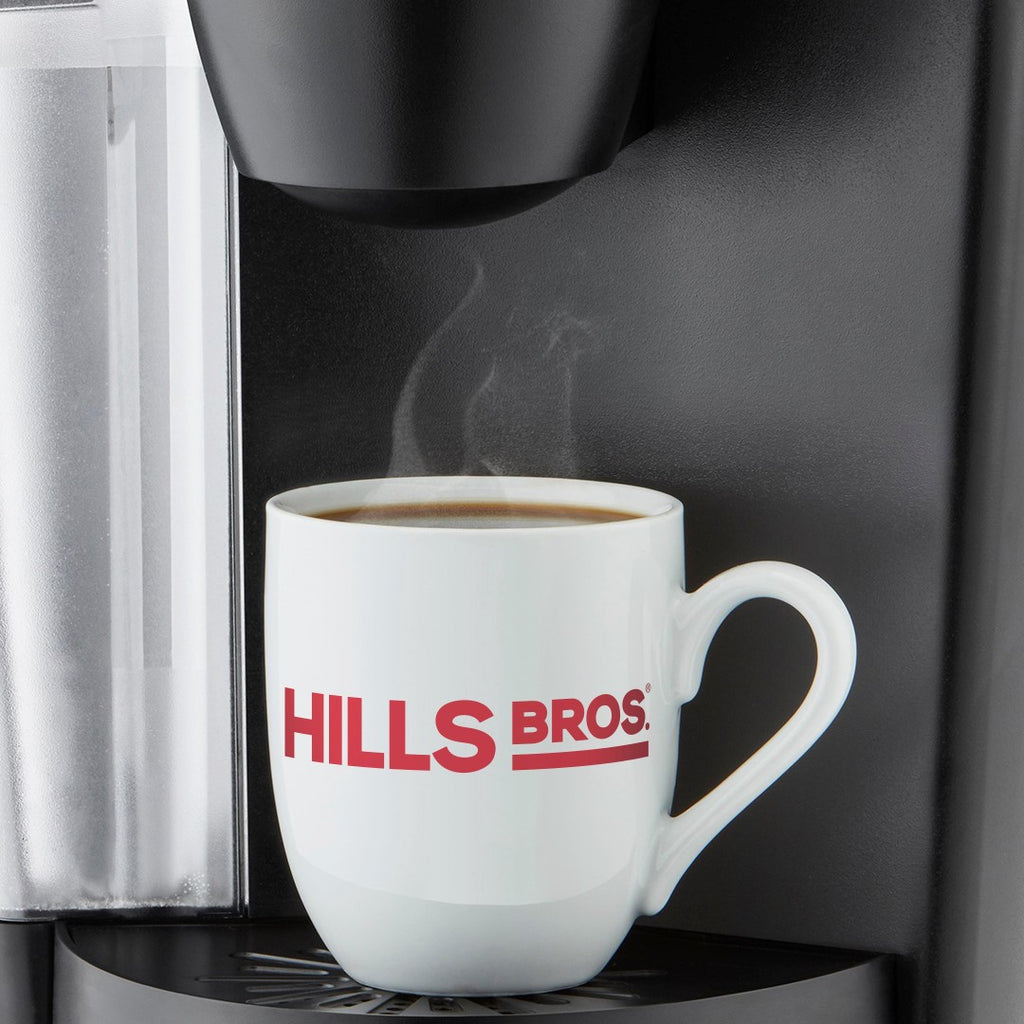 A white mug with "HILLS BROS." in red text sits under a coffee machine spout, with steam rising from the freshly brewed Original Blend - Medium Roast - Ground made from premium Hills Bros. Coffee beans inside.