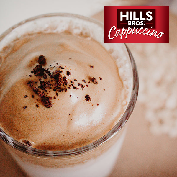 A glass of cappuccino topped with frothy milk and sprinkled with coffee grounds. The Hills Bros. Cappuccino logo is visible in the upper right corner.