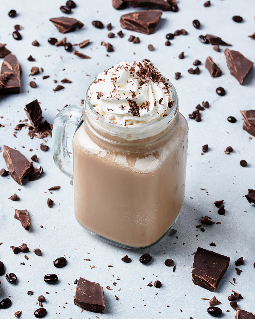 A mason jar filled with a Hills Bros. Frappes Chocolate Espresso frappé, topped with whipped cream and chocolate shavings, surrounded by coffee beans and chocolate pieces on a light surface.