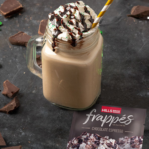 A glass jar filled with chocolate espresso frappé topped with whipped cream and chocolate drizzle on a black surface, surrounded by chocolate pieces and a Hills Bros. frappés product packet.