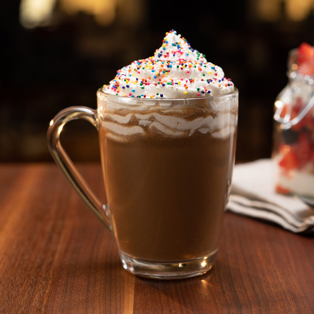 A glass mug of hot chocolate topped with whipped cream and colorful sprinkles, served on a wooden table alongside a packet of Hills Bros. Fat-Free French Vanilla - Instant Cappuccino Mix.