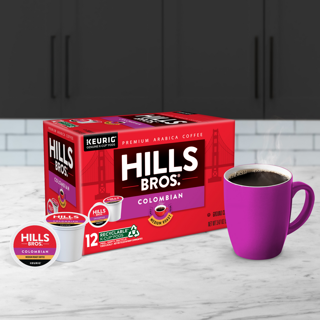 A box of Hills Bros. Coffee Colombian - Medium Roast - K-Cup® Pods sits on a marble countertop next to a pink mug filled with rich Arabica bean goodness.