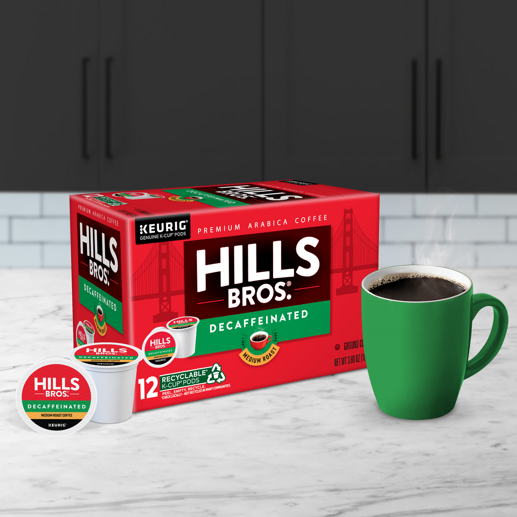 A box of Hills Bros. Coffee Decaf Blend Medium Roast Keurig K-Cup® Pods, crafted from premium Arabica beans, sits on the kitchen counter beside a steaming green mug filled with rich, flavorful coffee.