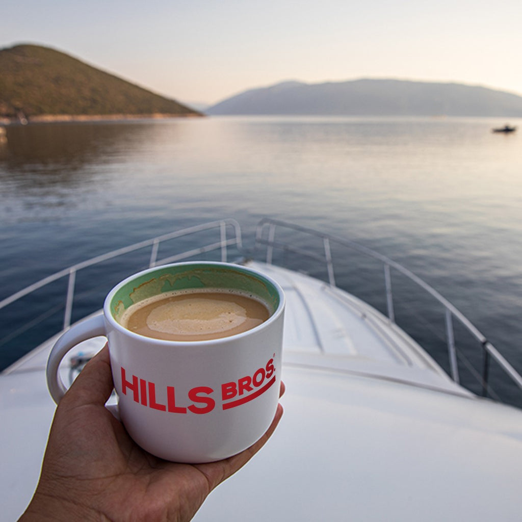 A hand holding a Hills Bros. Coffee mug, filled with 100% Colombian - Dark Roast - Ground, on the bow of a boat, with calm water and distant hills in the background.