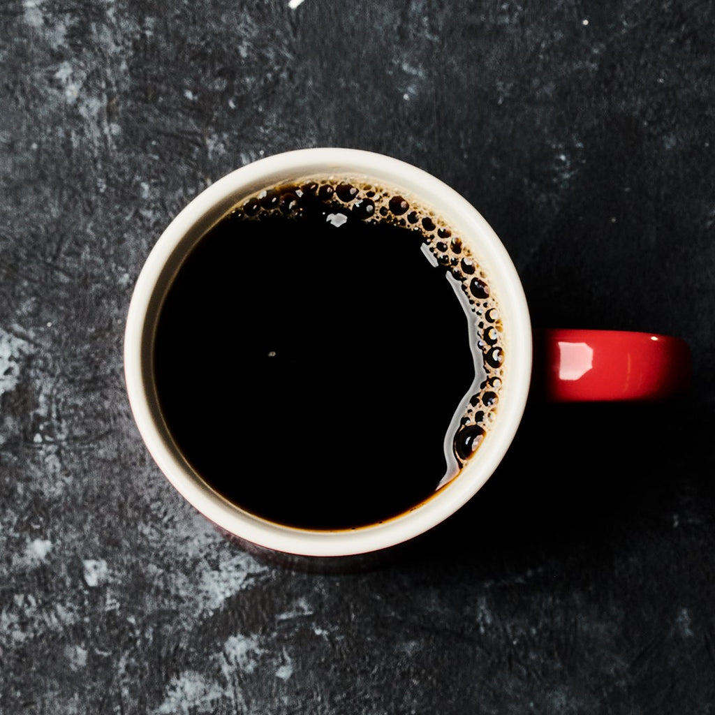 A red coffee mug filled with black coffee made from Hills Bros. Coffee Donut Shop - Medium Roast - Whole Bean - Premium Arabica is placed on a textured dark surface. The coffee shows slight bubbles on its surface.