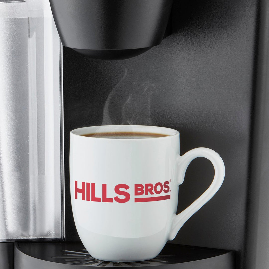 A white mug with the "Hills Bros. Coffee" logo placed under a coffee machine, filled with steaming coffee brewed from the Original Blend Medium Roast ground coffee.