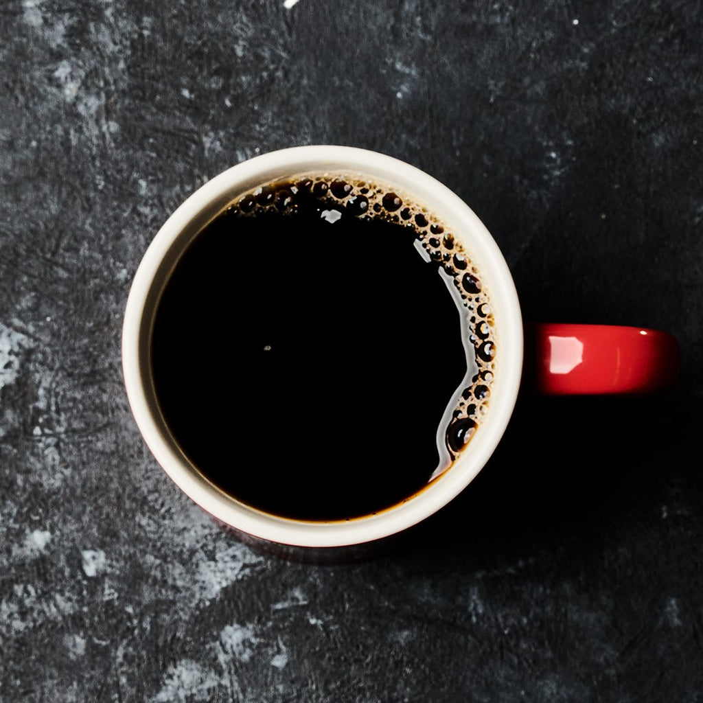 A red mug filled with steaming black coffee, brewed from Hills Bros. Coffee Original Blend - Medium Roast - Ground, sits on a textured dark surface.