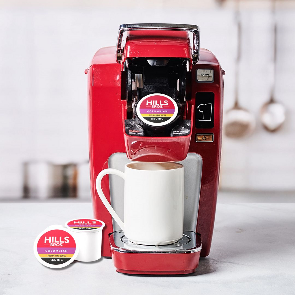 A red single-serve coffee maker with a "Hills Bros. Coffee" Colombian - Medium Roast - K-Cup® pod inside is on the countertop. A white mug waits under the dispenser, while another Arabica beans K-Cup® pod lies nearby.