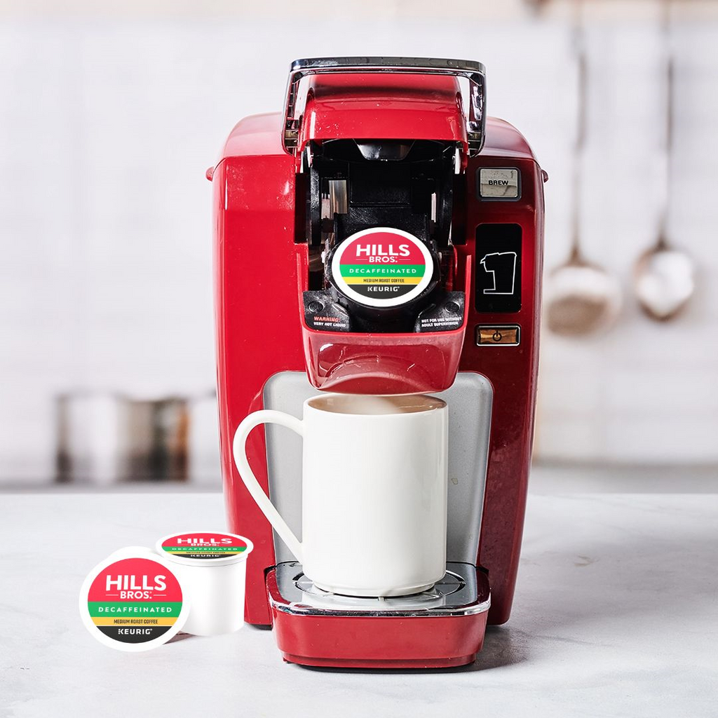 A red single-serve coffee maker with a white mug beneath the spout has a "Hills Bros. Coffee" K-Cup® pod labeled "Decaf Blend - Medium Roast" inserted. An additional pod rests on the counter nearby.