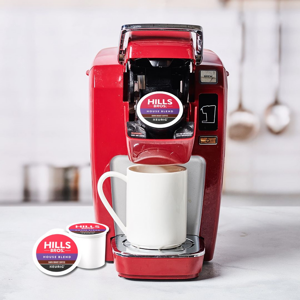 A red single-serve coffee maker is placed on a countertop with a white mug beneath its spout. Beside it are two K-Cup® pods labeled "House Blend - Dark Roast" by Hills Bros. Coffee, guaranteeing a rich taste from premium Arabica beans.