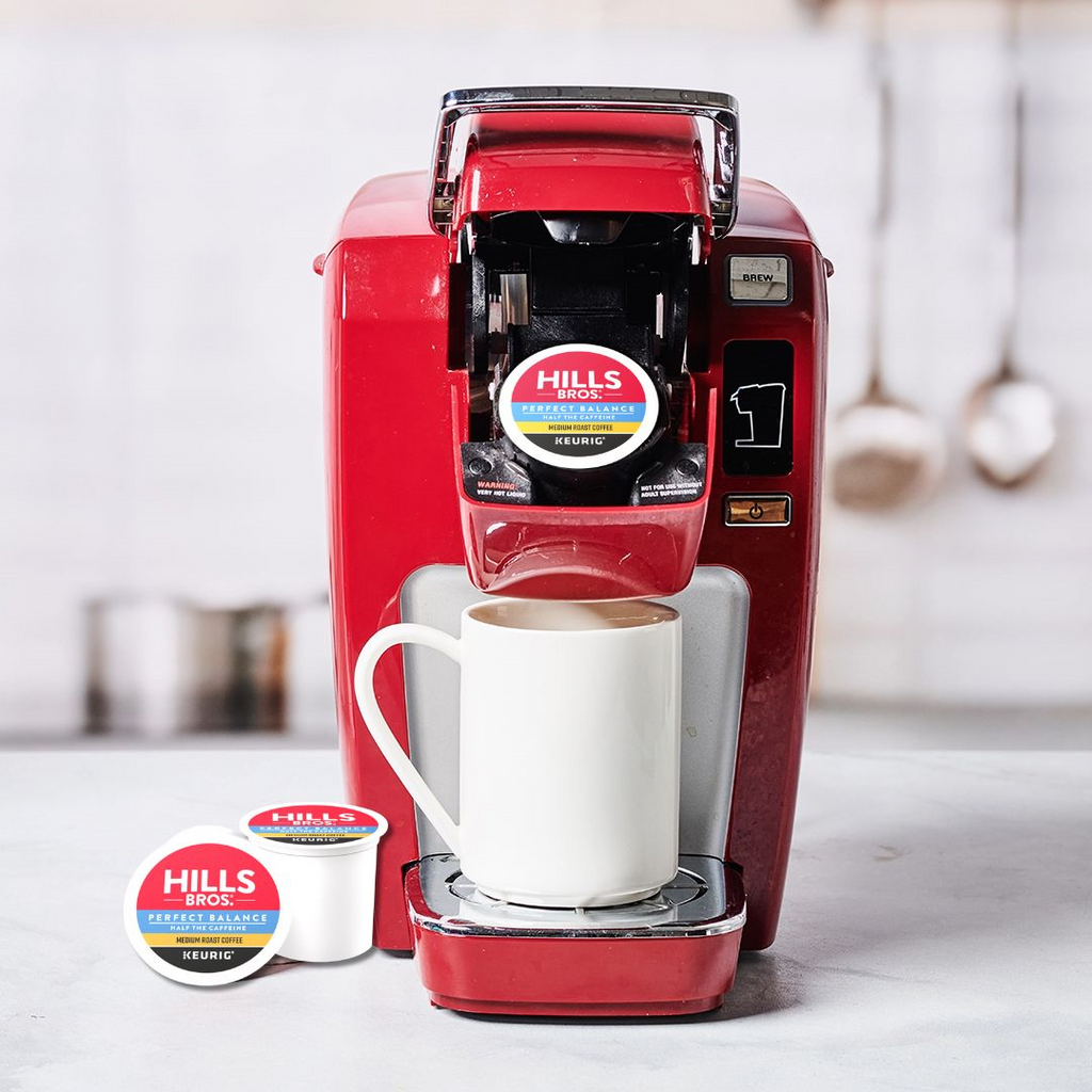 A red single-serve coffee maker stands with a white mug, containing a Hills Bros. Coffee Perfect Balance Medium Roast K-Cup® Pod, crafted from premium Arabica beans. Two more pods are neatly placed beside it on the counter, ready to provide more rich flavors.