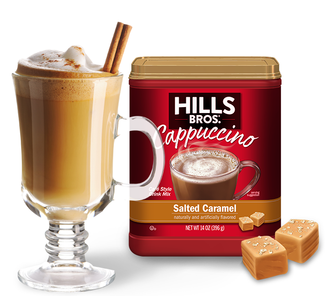 A glass mug of cappuccino with a cinnamon stick next to a 14-ounce container of Hills Bros. Salted Caramel Cappuccino mix and two pieces of caramel candy.
