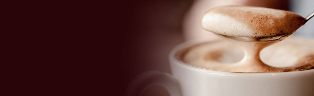 Close-up of a spoon lifting a layer of frothy milk from a cup of cappuccino, showing the creamy texture against a blurred background.